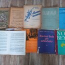 Fotografie zobrazuje deset různých tištěných hudebních notových sešitů a učebnic rozložených na dřevěné podlaze. Horní řada obsahuje sešity s názvy „…