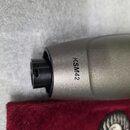Detail spodní části šedého mikrofonu s matným povrchem. Na těle je černý nápis KSM42. Mikrofon leží na červeném sametovém podkladu v šedé pěnové výpl…