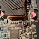 Detailní záběr na elektronickou základovou desku s komponenty. V horní části je černý hliníkový chladič s měděným základem. Na desce jsou viditelné č…