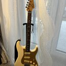 Elektrická kytara s bílým tělem, hnědým pickguardem a světlou dřevěnou hlavou. Na hlavě je viditelné logo Fender. Kytara má tři snímače, pět bílých l…