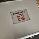 Stříbrný transportní kufr z hliníku. Uprostřed víka je připevněna čtvercová plaketa s nápisem SE ELECTRONICS a červeným logem SE. Povrch kufrů vykazu…