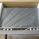 Šedý elektronický přístroj v ochranné plastové fólii, umístěný v přepravním kartonu s bílým polystyrenem. Na přední straně jsou vidět kovové ovládací…