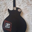 Zadní strana černé elektrické kytary s bílým lemováním. Na těle je nalepeno hnědé balení strun s logem a nápisem DUNLOP STRINGS. V horní části je vid…
