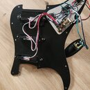 Černý pickguard s instalovanou elektronikou a třemi snímači. Na snímačích jsou viditelné bílé štítky s nečitelným textem. K elektronice vedou červené…