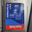 Šedý plastový adaptér s modrou a červenou nálepkou. Na nálepce je bílý nápis 6-in-1 PC Card Adapter a červené logo SanDisk. Jsou zde rovněž viditelné…
