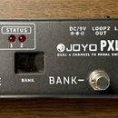 Černý efektový spínací systém s nápisem JOYO PXL-LIVE DUAL 4 CHANNEL FX PEDAL SWITCHING SYSTEM. Zařízení má tři kovové spínače označené MUTE, BANK- a…