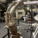 Detailní pohled na hradek a horní část saxofonu v kovovém provedení. Povrch nástroje vykazuje viditelné opotřebení, škrábance a známky koroze. Na nás…