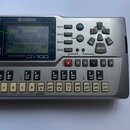 Stříbrný sekvencer s displejem a množstvím tlačítek. Na horní straně je logo YAMAHA a pod displejem nápis MUSIC SEQUENCER QY100. Zařízení vykazuje mí…