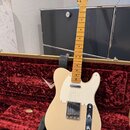 Světle béžová elektrická kytara typu Telecaster s bílým pickguardem, umístěná v otevřeném kufru s červeným sametovým vnitřkem. Na hlavě hnedku je vid…