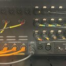 Zadní strana černého rackového audio zařízení s mnoha zapojenými kabely. Jsou zde viditelné nápisy OUT, POWER, CONTROL/AUDIO NETWORK, AES50 SUPERMAC…