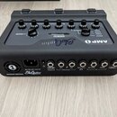 Černý kytarový procesor s nápisem GluGuitar AMP 1. Na horní straně jsou umístěny ovládací knoflíky a přepínač s nápisy MASTER, OVERDRIVE a různé reži…
