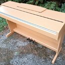 Světle hnědé digitální piano v dřevěném provedení s klaviaturním krytem. Nástroj má tři černé pedály v dolní části. Na povrchu nejsou viditelné žádné…