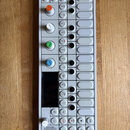 Bílý syntezátor OP-1 s řadou ovládacích prvků a displejem. Zařízení obsahuje čtyři barevné otočné knoflíky (oranžový, bílý, zelený a modrý) a několik…