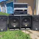 Sada zvukové techniky složená ze čtyř černých reproduktorů s kovovými mřížkami a dvou rackových zesilovačů. Na reproduktorech je vidět bílé logo s pí…