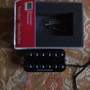Černý humbucker s dvěma řadami šesti ladících šroubů. Na spodní části je bílý nápis Seymour Duncan. Vedle nástroje je otevřená krabička s červeným št…