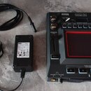 Černý sampler Korg Kaoss Pad Dynamic Effects Sampler s červeně podsvíceným displejem. K zařízení patří černý napájecí adaptér s nápisem KORG a dva če…