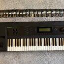 Černý klavír s nápisy KURZWEIL a K2000 V2. Vedle nástroje je položen manuál s názvem K2000 MUSICIAN'S GUIDE a KURZWEIL, spolu s elektronickou deskou.…