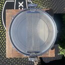 Fotografie zachycuje pohled z horní strany do vnitřku kovového bubnu, pravděpodobně malého snare drumu. Uprostřed je vidět napnuté bílé blána a přes…