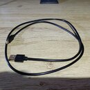 Černý USB kabel s konektory USB-A a Micro-USB. Kabel je složen do kroužku a položen na světlém dřevěném povrchu. Na kabelu nejsou viditelné žádné zna…