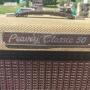 Detail kytarového komba potaženého béžovou texturou s hnědým koženým úchem. Na přední straně je kovový štítek s nápisem Peavey Classic 50. Povrch ští…