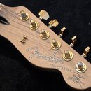 Detail hlavy kytary z přírodního dřeva se zlatými ladičkami. Na dřevě je bílý nápis Fender a pod ním v uvozovkách text "TELECASTER". V pravém dolním…