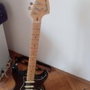 Černá elektrická kytara typu Stratocaster s jasným dřevěným hředníkem a krčkem. Na hlavě je vidět nápis Fender STRATOCASTER. Kytara má tři snímače, b…