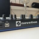 Zadní strana černého elektronického zařízení s modrým spodním okrajem. Na zadním panelu je bílý nápis novation s doprovodným logem, vstup pro napájen…
