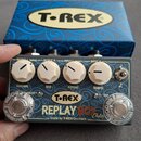 Kytarový efekt s nápisem T-REX REPLAY BOX Delay a doplňkem Made by T-REX Denmark. Zařízení má šedý povrch, čtyři bílé ovládací knoflíky s popisky VOL…