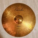 Zlatavý kovový činel s kovaným povrchem. Na horní části je černý nápis Paiste a ve spodní části text Dimensions Hand Hammered Ride. Povrch vykazuje d…