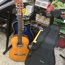 Na fotografii jsou dvě akustické kytary a černý pouzdro typu gig bag. Přední kytara je klasická s přírodním dřevem a v otvaru má ozdobný prstenec, za…