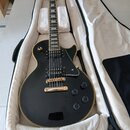 Černá elektrická kytara s tvarem Les Paul, zlatým hardwarem a bílým lemováním těla. Nástroj je umístěn v otevřeném plišovém kufru bílé barvy. Na hlav…