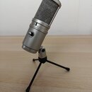 Stříbrný kondenzátorový mikrofon s kovovou mřížkou, umístěný na černém trojnožném stativu. Na těle mikrofonu je nápis SuperLux a ovládací prvek s ozn…