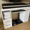 Bílá pracovní plocha s přívěsem na klávesnici, na které je umístěn černý digitální klavír. Pod deskou jsou dvě bílé šupláky s nápisy URBAN-X, vysuvac…