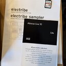 Na bílém papíře jsou nápisy KORG, electribe MUSIC PRODUCTION STATION a electribe sampler MUSIC PRODUCTION. Vedle papíru leží malá SD karta značky PHI…