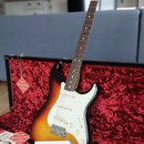 Elektrická kytara s tónem sunburst a bílým pickguardem v otevřeném kufru s červeným vnitřkem. Na hlavě hředu je logo Fender. Kytara je doplněna o cer…