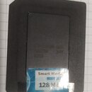 Černá paměťová karta SmartMedia s modrobílou nálepkou. Na nálepce je nápis Smart Media a 128 MB. V horní části těla jsou vyražená písma a čísla: 70NS…