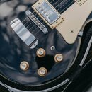Černá elektrická kytara s bílým pickguardem a dvěma humbuckery s kovovými kryty. Na kytáře jsou čtyři zlaté otočné knoflíky s nápisy VOLUME, VOLUME,…