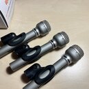 Tři stříbrné dynamické mikrofony s kovovými mřížkami a černými plastovými držáky. Na tělech mikrofonů jsou viditelné rýhované prstence, ale žádná zna…