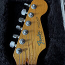 Zadní strana hlavy kytary z přírodního dřeva s šesti stříbrnými ladičkami. Na dřevě je černý nápis STRATOCASTER MADE IN U.S.A. a logo Fender s označe…