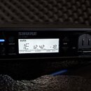Černý přijímač s nápisem SHURE GLXD4+. Displej zobrazuje text „auto“ a hodnoty: group 1, channel E, battery 12 hr 42 mn, audio gain + 18 dB. Na panel…