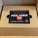 Černý napájecí adaptér pro efektové pedály v kartonové krabici. Na horní straně je bílý a červený nápis VOODOO LAB PEDAL POWER X4 a text HYBRID DC TR…