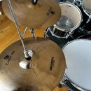 Dva mosazné činely na stojanu s nápisy CAST38 a 14" HIHAT-BOTTOM BY SONOR. V pozadí jsou vidět části bubnové sady značky SONOR v černém provedení. Po…