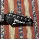 Černá hlava elektrické kytary s nápisem Ibanez a menším nápisem JS Series. Na hlavě jsou umístěny šest kovových ladiček. Povrch je lesklý a v dobrém…