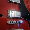 Detail červené elektrické kytary s dvěma humbuckery v kovových krytech a černým plechtrádem. Nástroj má chromovaný kobylník a strunení. Na povrchu la…