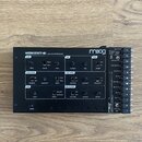 Černý analogový syntetizátor s nápisem WERKSTATT-01 ANALOG SYNTHESIZER a logem moog. Přístroj obsahuje různé ovládací knofíky, přepínače a sekce ozna…