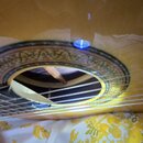 Detailní záběr na rozetu akustického nástroje s ozdobným ornamentem v zelené a červené barvě. V horní části rozety je umístěn malý modrý LED indikáto…