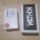 Na fotografii jsou dvě krabičky od napájecích zdrojů položené na dřevěném povrchu. Menší bílá krabička nese nápisy Rock Power® by Warwick®, Power Sup…
