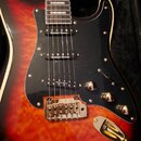 Elektrická kytara s červeně-žlutým gradientem a černým pickguardem. Na všech třech snímačích je bílý nápis Seymour Duncan. Kytara má tři kovové ovlád…