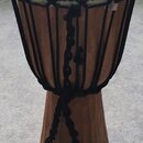 Dřevěný djembe s bílou membránou a černým provázkem. Na membráně je viditelné logo s písmenem B a nápisem MEINL. Na horním okrahu korpusu je nalepená…