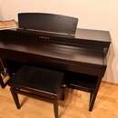 Tmavě hnědý digitální piano v dřevěném provedení s přívodním stolem na noty. Na přední straně je bílý nápis YAMAHA. Součástí je ladící stolek s černý…